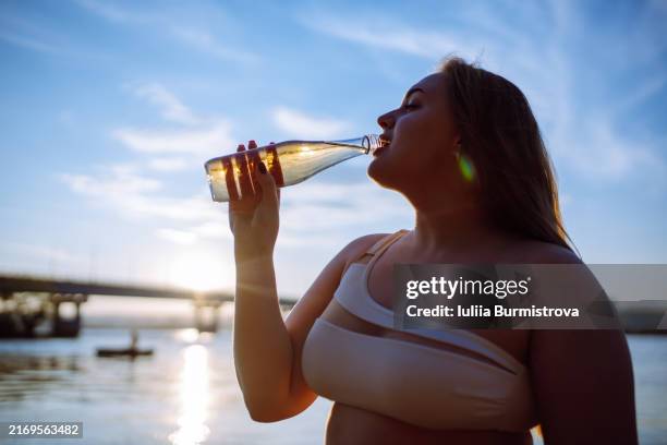 charming young woman enjoying refreshing sip of water from glass bottle on beach - junge frau allein stock-fotos und bilder