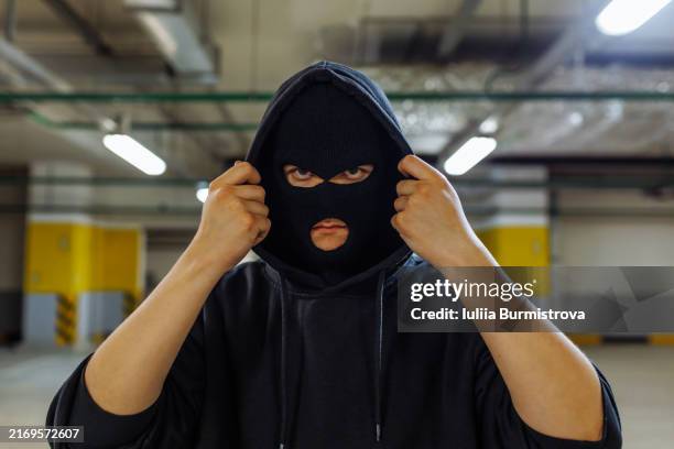 sinister-looking young man wearing face mask and hooded sweatshirt standing in empty parking garage - voleur photos et images de collection