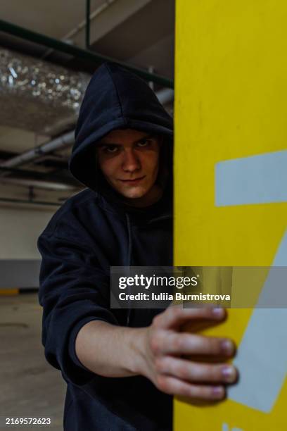 ominous looking young man peeking over corner in underground garage looking at camera sullenly - bandit stock pictures, royalty-free photos & images