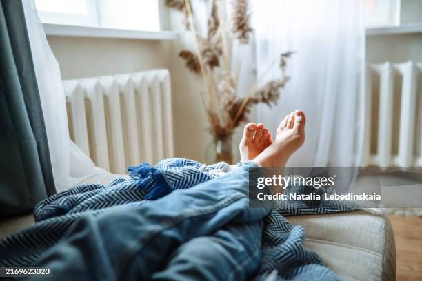 warmth and coziness at home: bare feet with a blanket on the couch next to the heating radiators. - laziness stock pictures, royalty-free photos & images