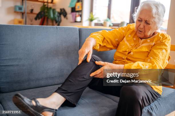 old woman feeling pain at home in living room while sitting on sofa bed - leg stock pictures, royalty-free photos & images