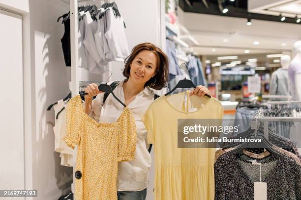 attractive woman holding two yellow dresses smiling at clothing store - habilleuse styliste photos et images de collection