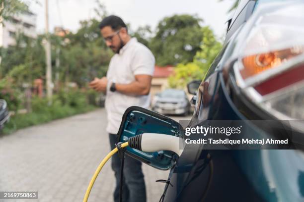 young man waiting near his ev while it's charging. - electric vehicle stock pictures, royalty-free photos & images