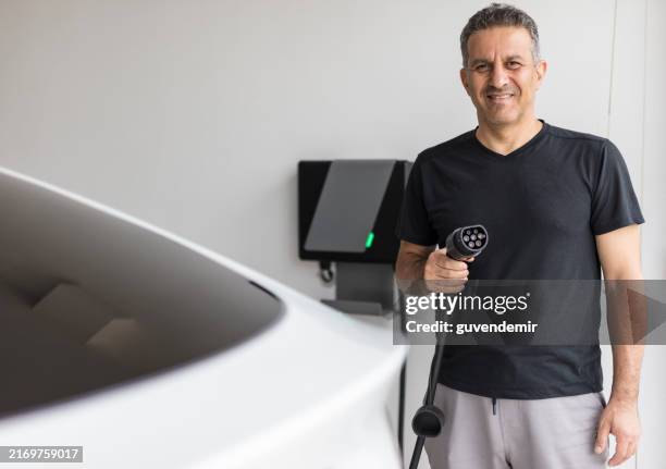 mature man holding electric plug to charge his electric car at home - battery charger stock pictures, royalty-free photos & images