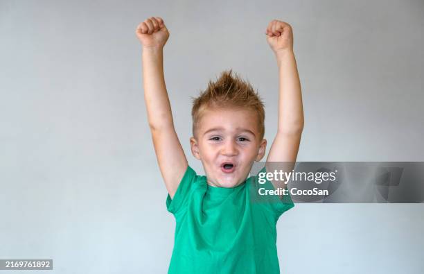 cheerful young boy with spiky hair in a green t-shirt raising his arms in excitement against a plain background. - only boys stock pictures, royalty-free photos & images