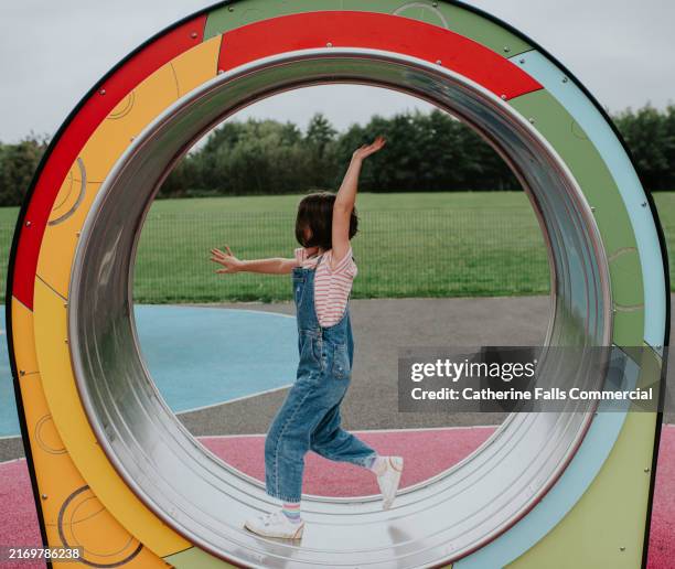 young girl joyfully playing inside a colourful playground hamster wheel structure on a cloudy day in a grassy park - draaimolen stockfoto's en -beelden