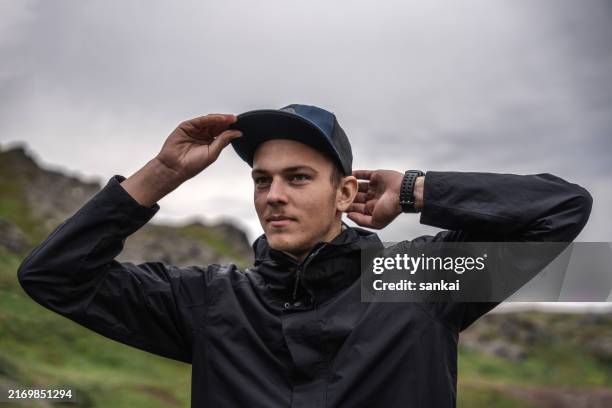 portrait of the young man adjusting his baseball cap in the mountains, dramatic scenery - paisagem espetacular imagens e fotografias de stock
