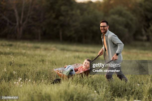 cheerful father pushing his son in wheelbarrow through grass. - kruiwagen stockfoto's en -beelden