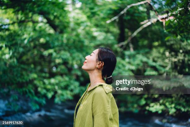 beautiful young asian woman meditating in the nature with her eyes closed, setting herself free and feeling relieved. enjoying fresh air and breathing in the calmness with head up. freedom in nature. connecting with nature. health and wellness concept - meditieren stock-fotos und bilder