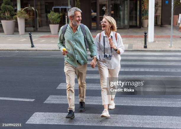 happy mature couple crossing street hand in hand on a sunny day - couple crossing street stock pictures, royalty-free photos & images