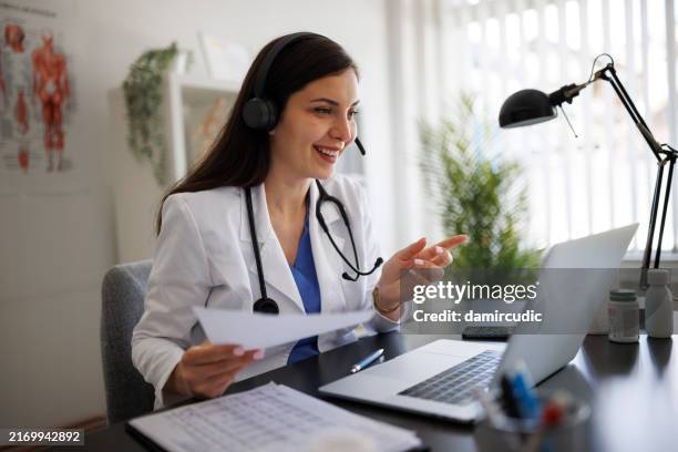 smiling female doctor in her office, using a laptop to give online consultations while wearing a headset - telemedicine stock pictures, royalty-free photos & images