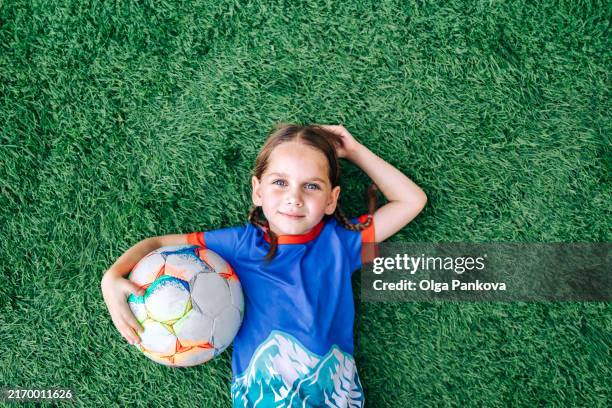 young girl, a child soccer player, lies hugging a ball on the field, football - tenue de football photos et images de collection