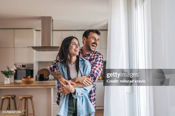junges fröhliches paar, das sich am fenster umarmt, im hintergrund ist die küche zu sehen. - glücklichsein stock-fotos und bilder