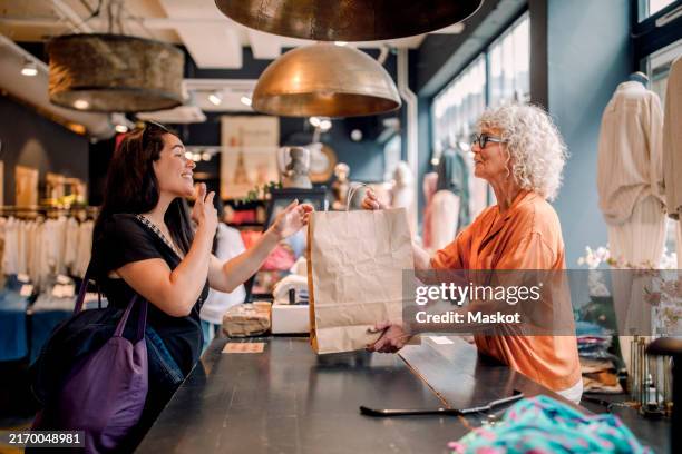 senior female owner giving bag to customer at checkout in clothing store - einkaufen stock-fotos und bilder
