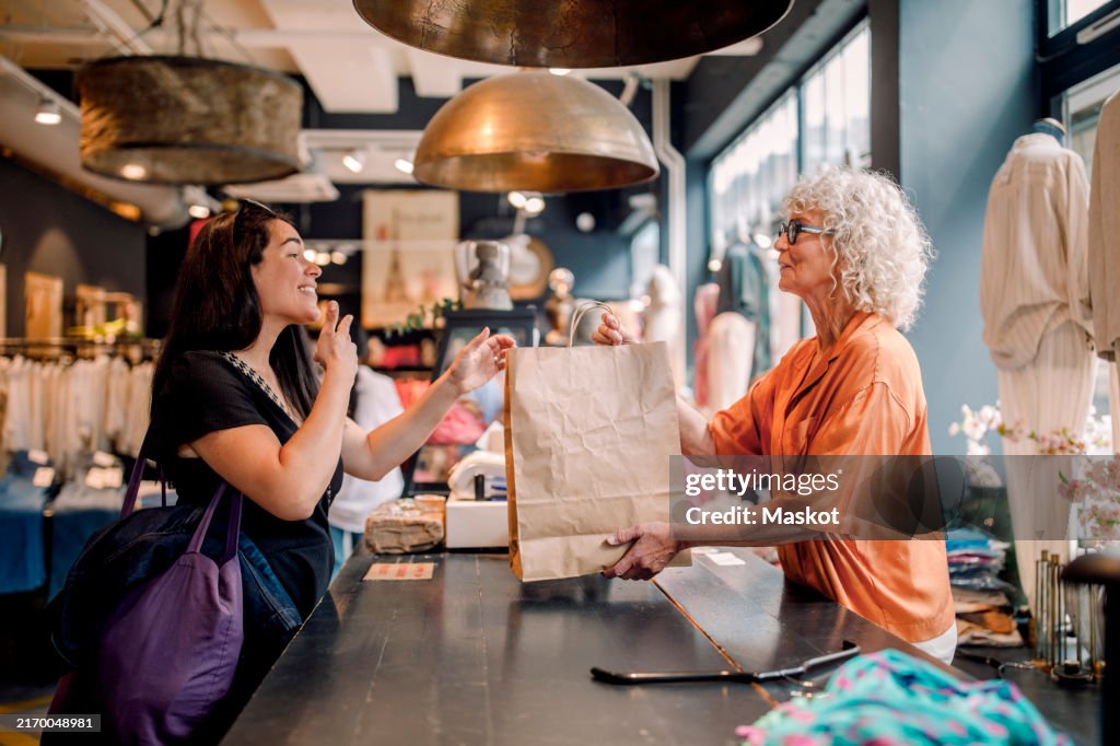 Senior female owner giving bag to customer at checkout in clothing store