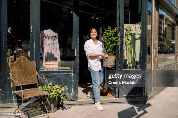 smiling young female entrepreneur holding plant while walking out of store - photo store stock pictures, royalty-free photos & images