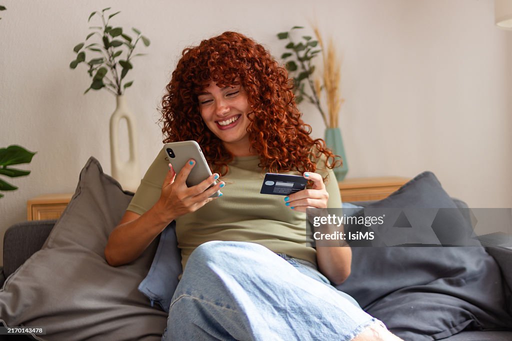 Happy woman using smartphone and credit card for online shopping on sofa at home