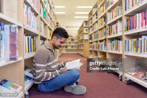 young man sitting on the floor and reading a book in library - sitting on ground stock pictures, royalty-free photos & images
