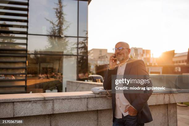 senior businessman talking on the phone in front of the office building - golden hour stock pictures, royalty-free photos & images