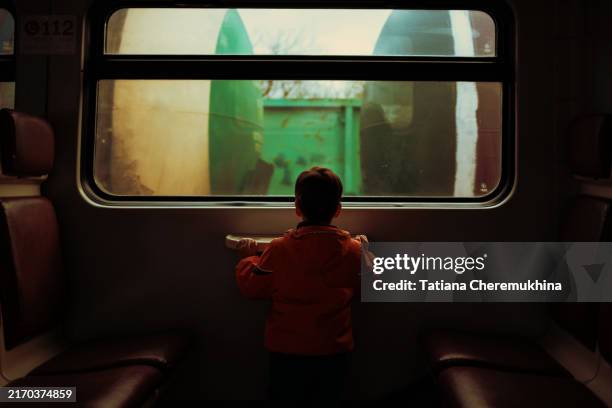 a child with his back to the camera looks out the window of a train. several rows of freight cars are visible outside the window. - russian boy stock pictures, royalty-free photos & images