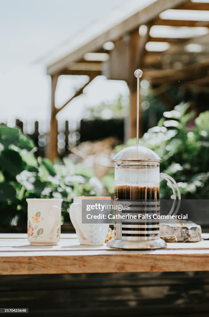 Coffee brewing with a French press on a sunny patio surrounded by lush greenery and two mugs ready for serving