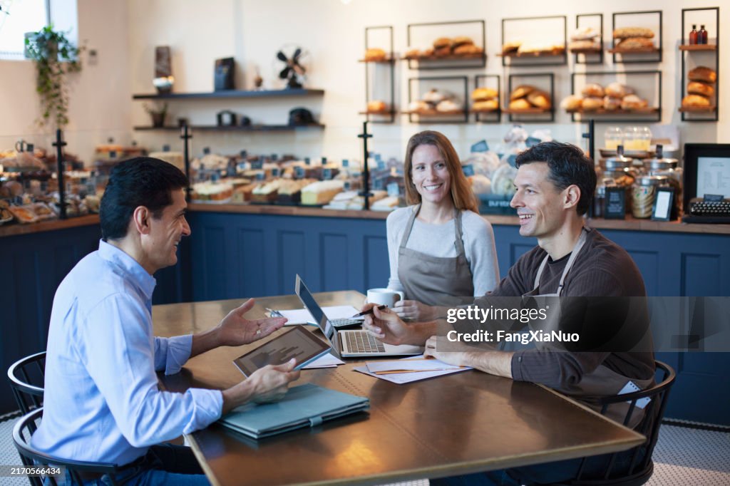 Mature Middle Eastern man financial advisor insurance agent talking advice with mature women and mature man owner staff employee sitting together in small business bakery cafe using laptop computer digital tablet planning