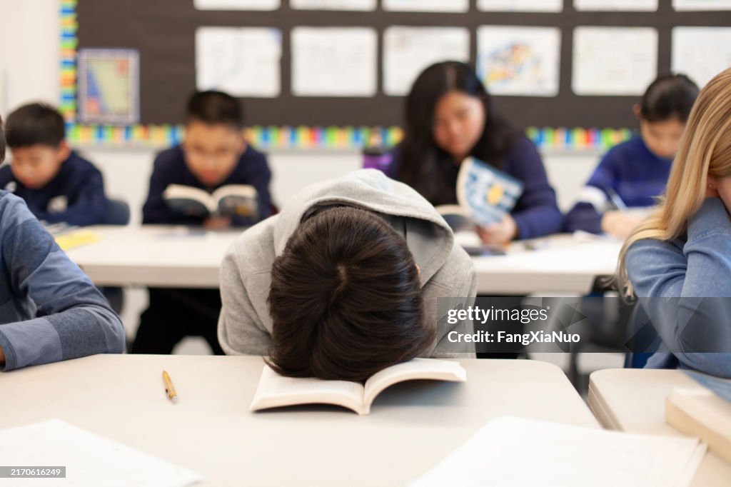 Elementary school junior high boy student sitting at desk in classroom with head in book in frustration sleeping tired exhaustion boredom