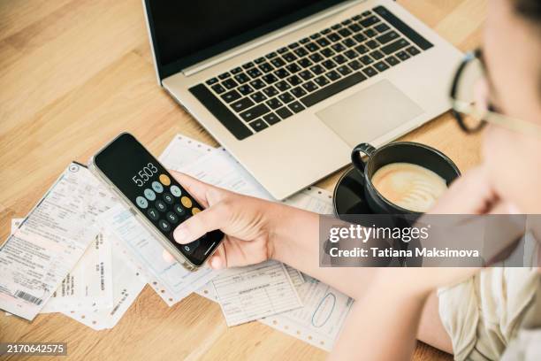 a woman calculates utility bills on a calculator on her phone. woman sits on the table with laptop and cup of cappuccino. - bank statement stock pictures, royalty-free photos & images