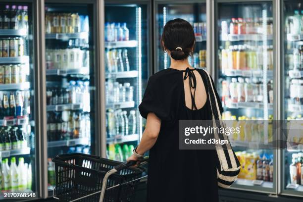 rear view of young asian woman with shopping cart, doing grocery shopping in supermarket. she is is standing in front of a row of freezers and choosing products. eating well. choosing quality and healthy source of food in environmentally conscious way - food allergy stock pictures, royalty-free photos & images