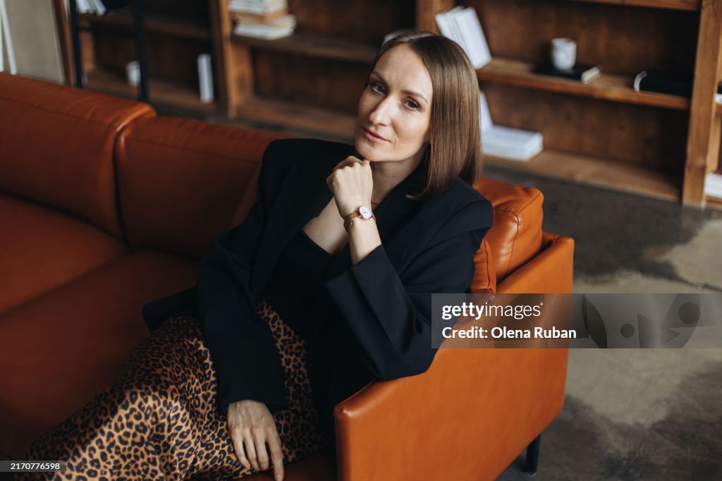 Young woman in a black jacket and a leopard skirt sitting on a couch.