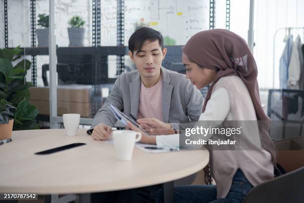 two colleagues sitting at a table during a meeting, discussing work while looking at a tablet device, with coffee cups nearby - performance review stock pictures, royalty-free photos & images