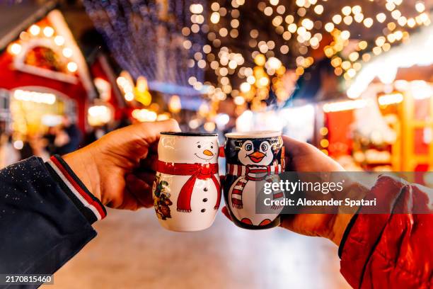 two persons drinking glühwein (mulled wine) from snowman and penguin shaped christmas market in berlin, germany - marché de noel photos et images de collection