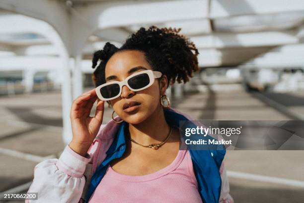 portrait of fashionable curly hair woman holding sunglasses at parking lot in sunlight - accessoires stock-fotos und bilder