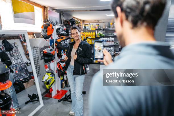 junger mann, der seine freundin fotografiert, während sie motorradjacken in einem motorradgeschäft anprobiert - motorradjacke stock-fotos und bilder