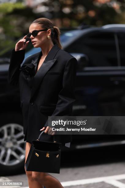Brooks Nader is seen wearing black cat-eye sunglasses from Dior, chunky gold hoop earrings, a black blazer with shoulder pads and a large black...