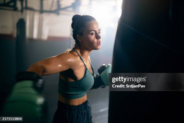 mujer joven, con guantes de boxeo, golpeando el saco de boxeo, mientras practica kickbox en el gimnasio - kick boxing fotografías e imágenes de stock