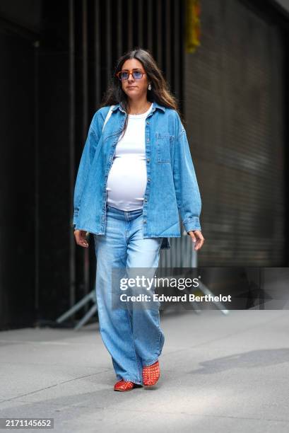 Guest wears red rimmed sunglasses with blue tinted lenses, silver earrings, white t-shirt, light blue oversized denim jean unbuttoned long sleeve...