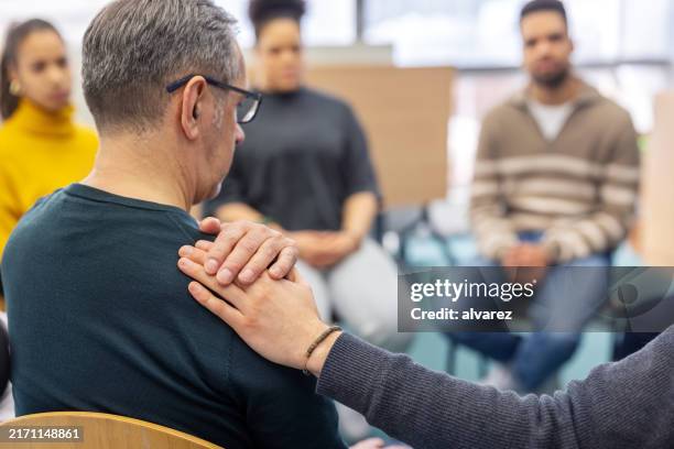 uomo anziano confortato da un altro partecipante alla terapia di gruppo al centro comunitario - malattia mentale foto e immagini stock