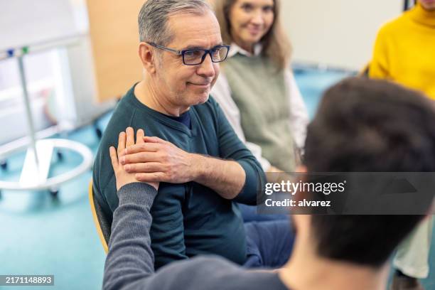 hombre mayor que recibe apoyo emocional de otro pasticante durante la sesión de terapia de grupo - terapia de grupo fotografías e imágenes de stock