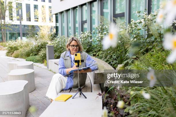 mature woman coach using smartphone to record vlog sitting outside near modern office building - tripod stock pictures, royalty-free photos & images