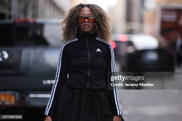 Fashion Week guest is seen wearing square black sunglasses with orange lenses, large gold hoop earrings, a cropped black zip jacket from Adidas with...