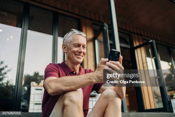 mature man uses a phone while sitting on a cozy sidewalk cafe in summer - baby boomer photos et images de collection