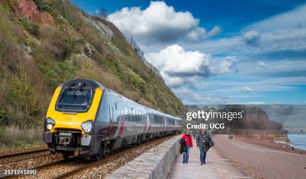 Train passes a middle-aged couple walking along the promenade on the seafront in Teignmouth, Devon, UK.