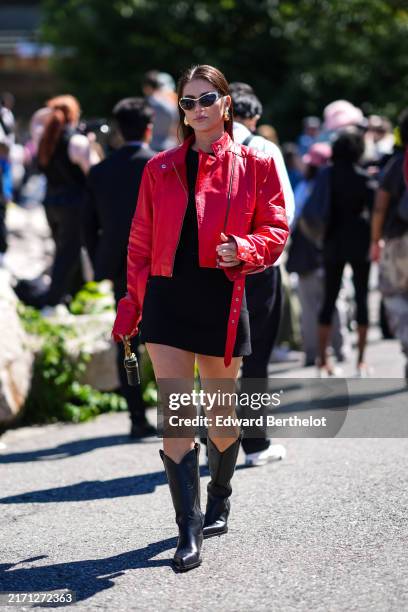 Guest wears black sunglasses, gold earrings, bright red cropped biker leather jacket, black mini dress, black mini leather bag, shiny black cowboy...