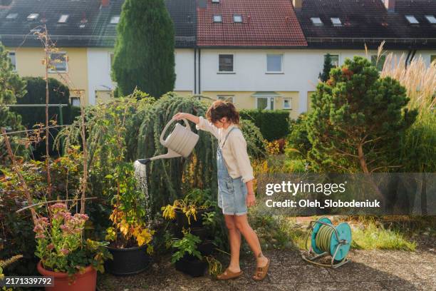 woman in denim overalls watering plants on her backyard - trädgård bildbanksfoton och bilder