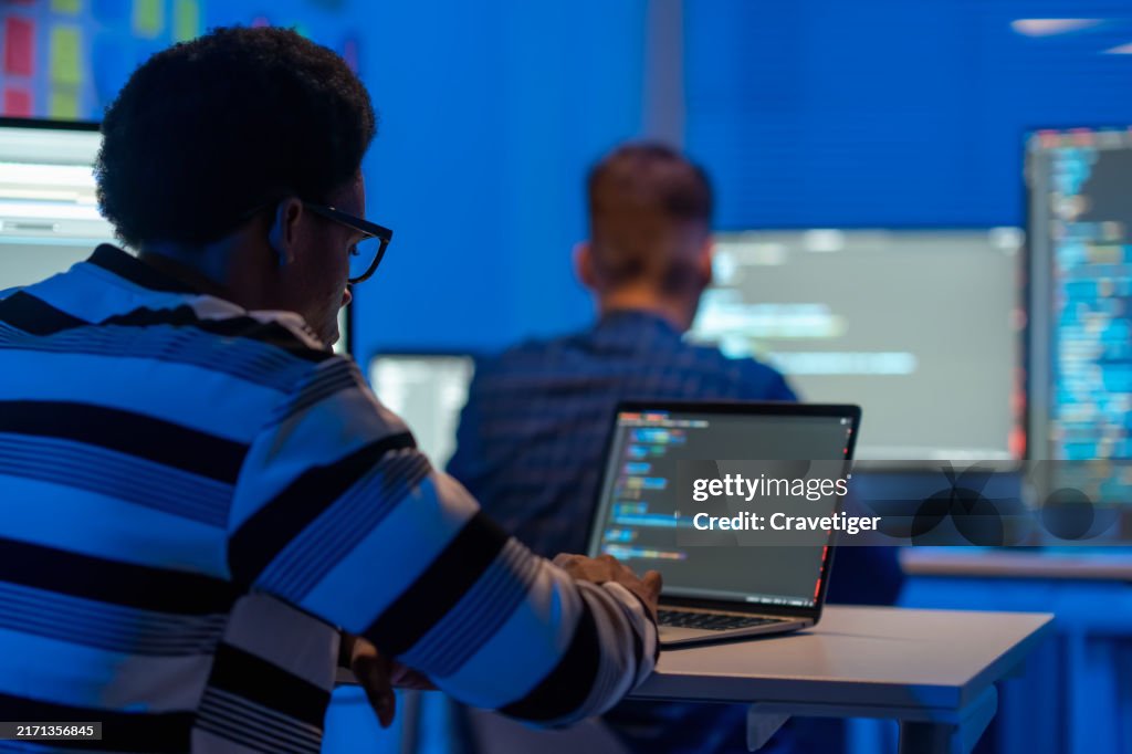 Rear view of coding programmer in Working on desktop computer for work remotely at data center room while his teamwork in the background. Programmer Creating code ,Modern Software, e-Commerce App Design , Programmer development concept.