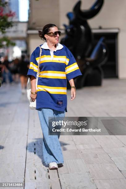 Guest wears dark brown sunglasses, white navy blue yellow striped short sleeve oversized shirt, white leather bag, a gold bracelet, a watch, gold...