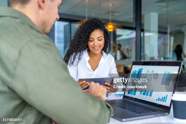 hombre de negocios y mujer de negocios en una reunión en la oficina. hay una computadora portátil sobre la mesa que muestra tablas y gráficos financieros y la mujer sostiene una tableta digital - panel de indicadores medios visuales fotografías e imágenes de stock