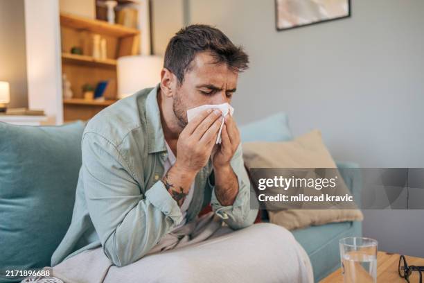 a sick mid adult man is sitting on a sofa. - erkältung und grippe stock-fotos und bilder
