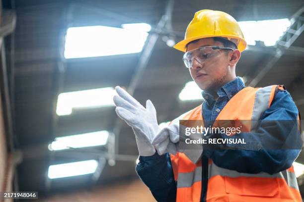 worker in yellow hard hat adjusts gloves and helmet in a hardware factory. - protective workwear stock pictures, royalty-free photos & images